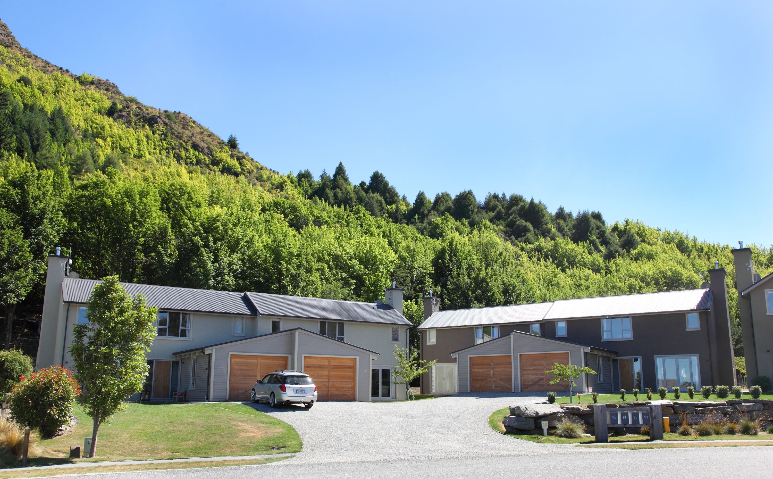 A modern residential area with two-story houses, a gravel driveway, and a forested hillside in the background.