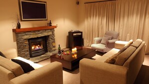 A living room with a stone fireplace, a flat-screen TV, a coffee table with candles, and a checkered armchair. - Arrowfield Apartments (Arrowtown)