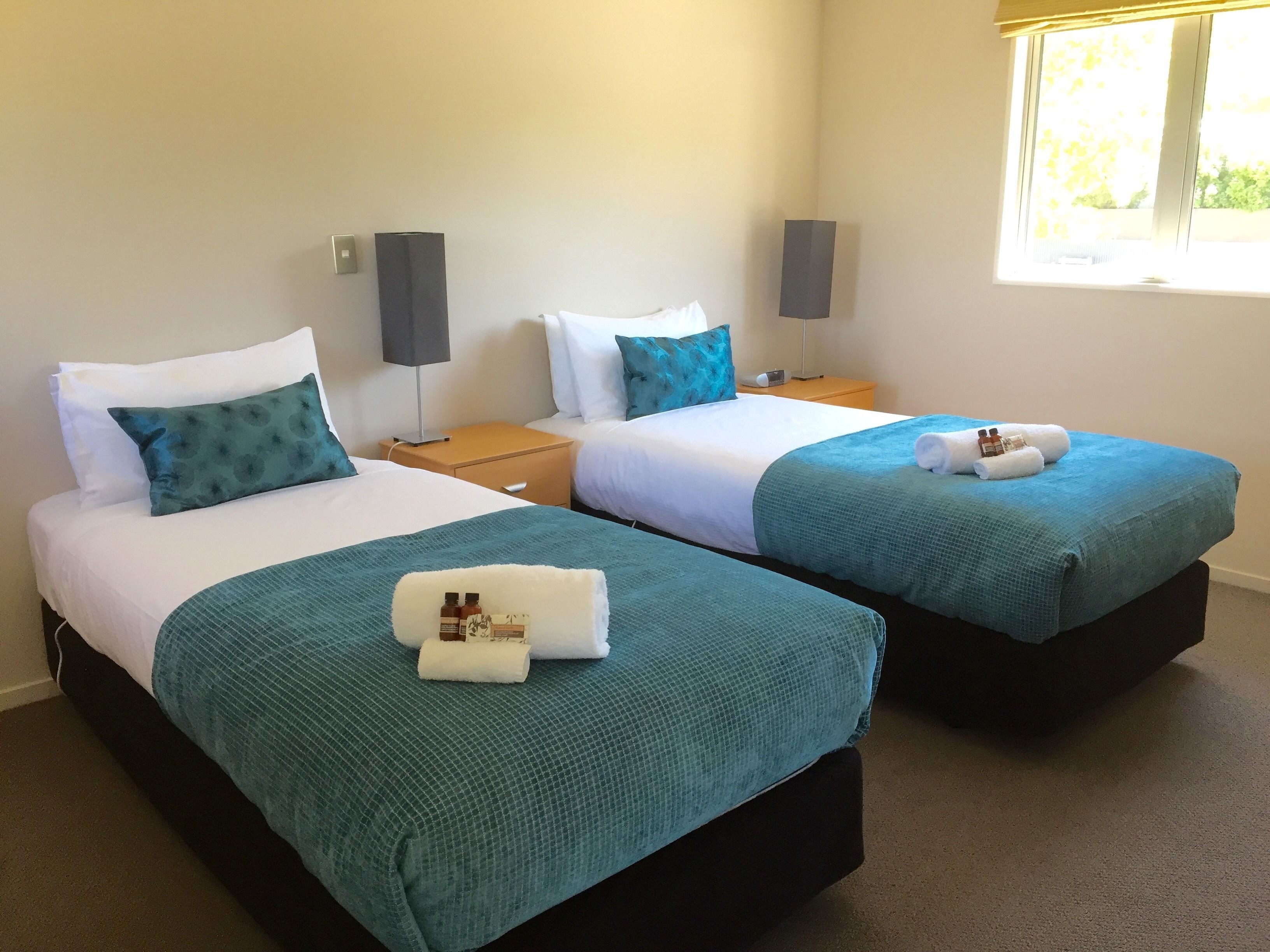 A hotel room with two single beds, each with a teal bedspread and white pillows. There are two bedside tables with lamps, a window with a view of greenery, and a carpeted floor.