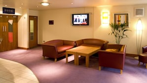 A hotel lobby with a reception desk, a television mounted on the wall, a seating area with a sofa and chairs, and a potted plant. - Premier Inn Heathrow Airport - M4/J4 (Hayes)