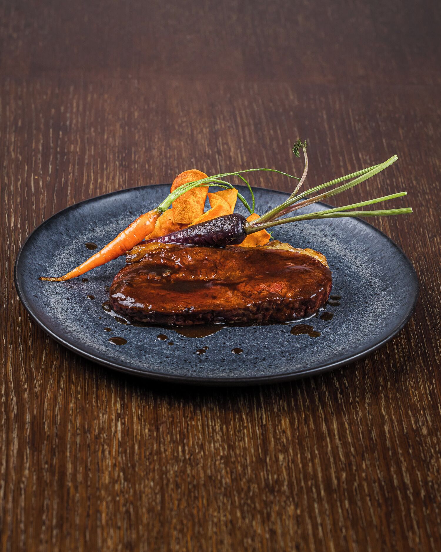 A plate with a steak, carrots, and a garnish on a dark plate.