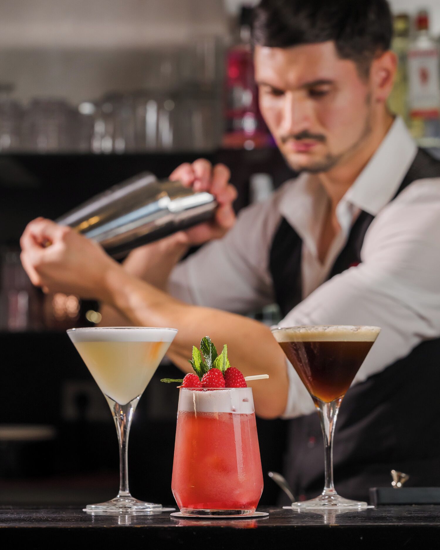 A bartender preparing drinks behind a bar with three different cocktails in front of him.