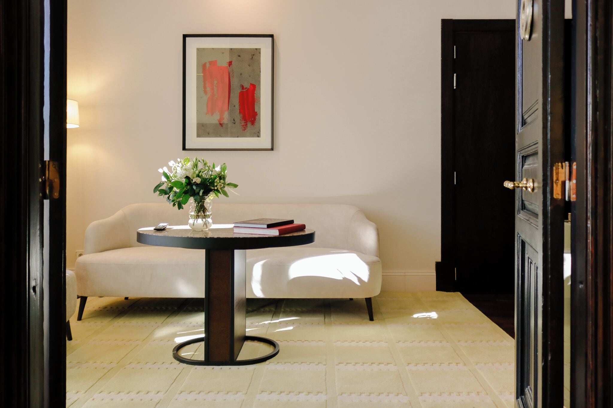 Una habitación con un sofá blanco, una mesa redonda con un jarrón de flores, un cuadro abstracto enmarcado y una puerta oscura.