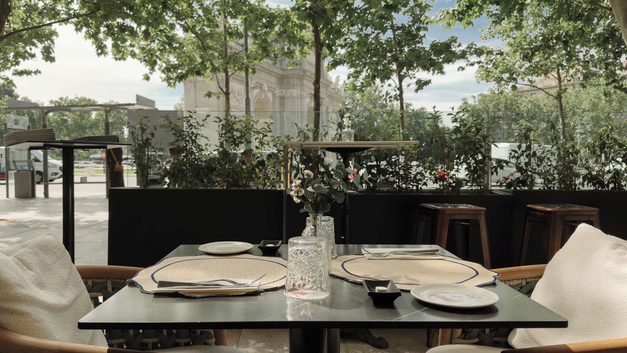 An outdoor dining area with a table set for two, surrounded by chairs and a view of a building in the background.