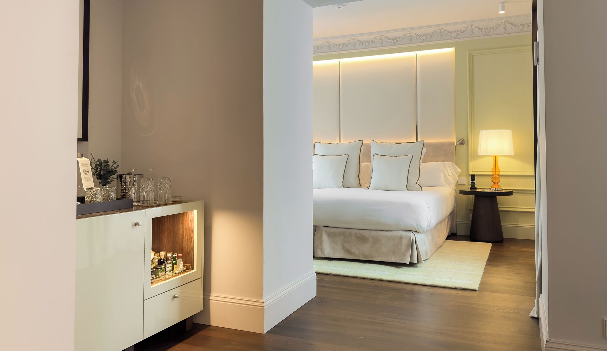 A hotel room with a bed, bedside table, lamp, and a vanity with a mirror and storage.