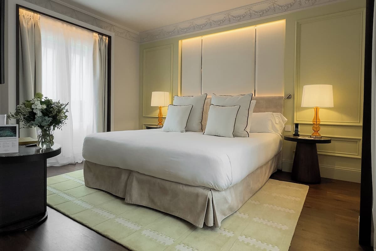 Een hotelkamer met een groot bed, twee nachtkastjes met lampen, een bureau met een bloevas en een groot raam met gordijnen.