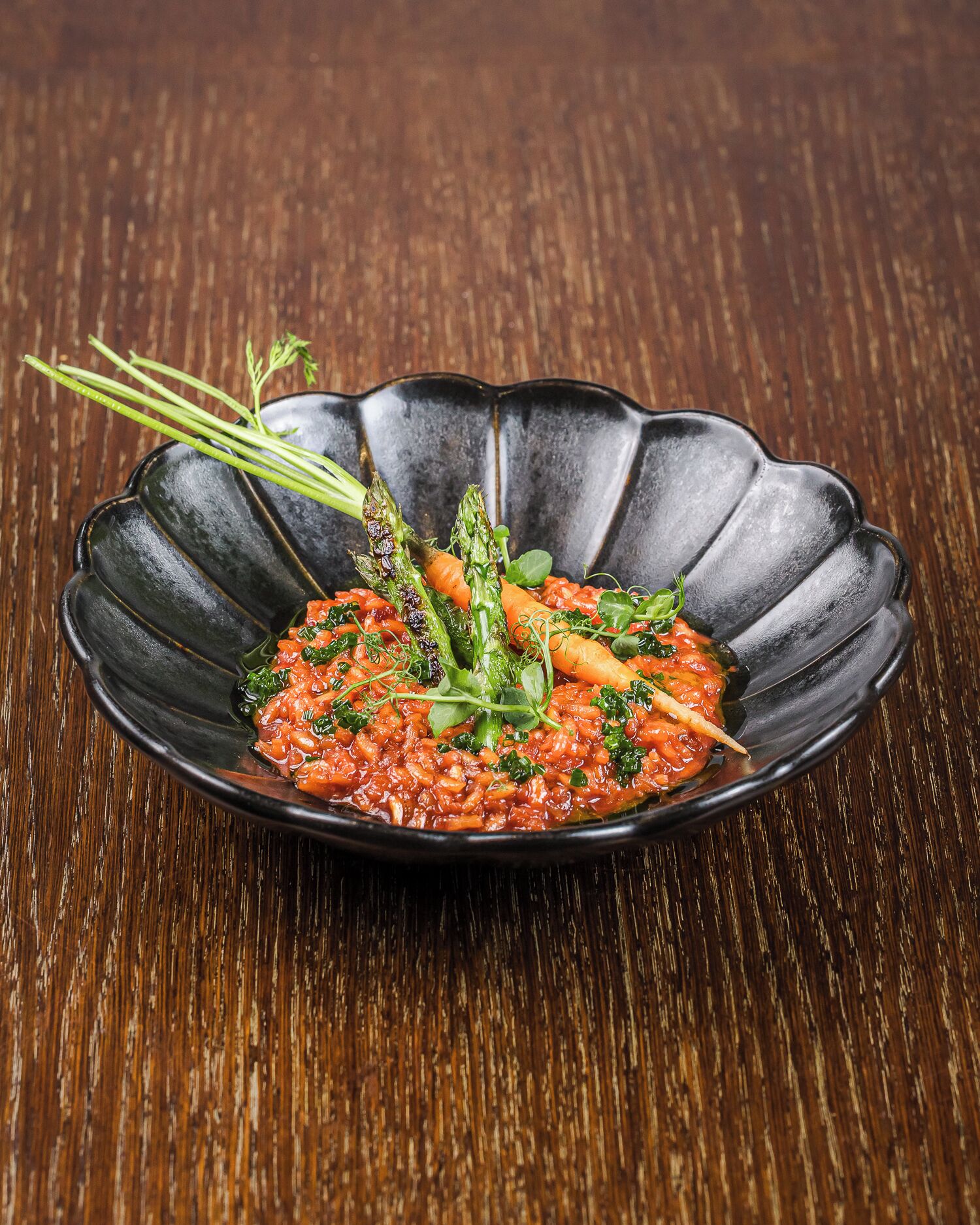 A black, scalloped-edge dish with a red sauce and vegetables on a wooden table.