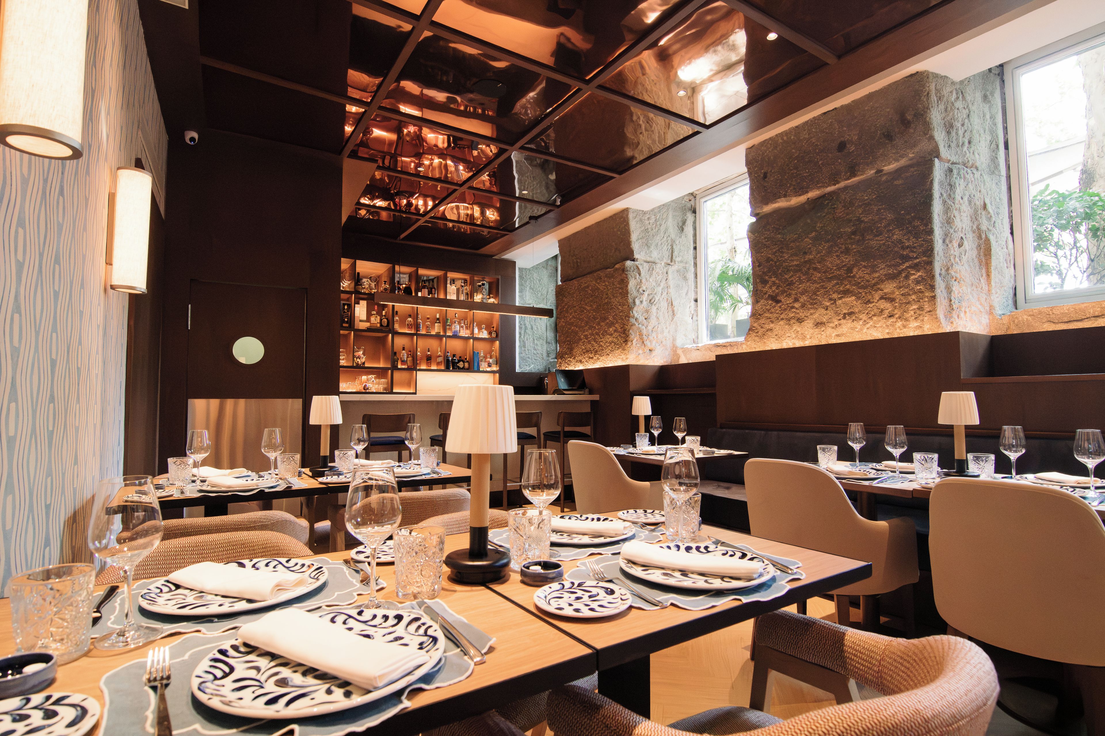 A restaurant interior with multiple dining tables set for guests, a bar area with shelves of bottles, and a stone accent wall.