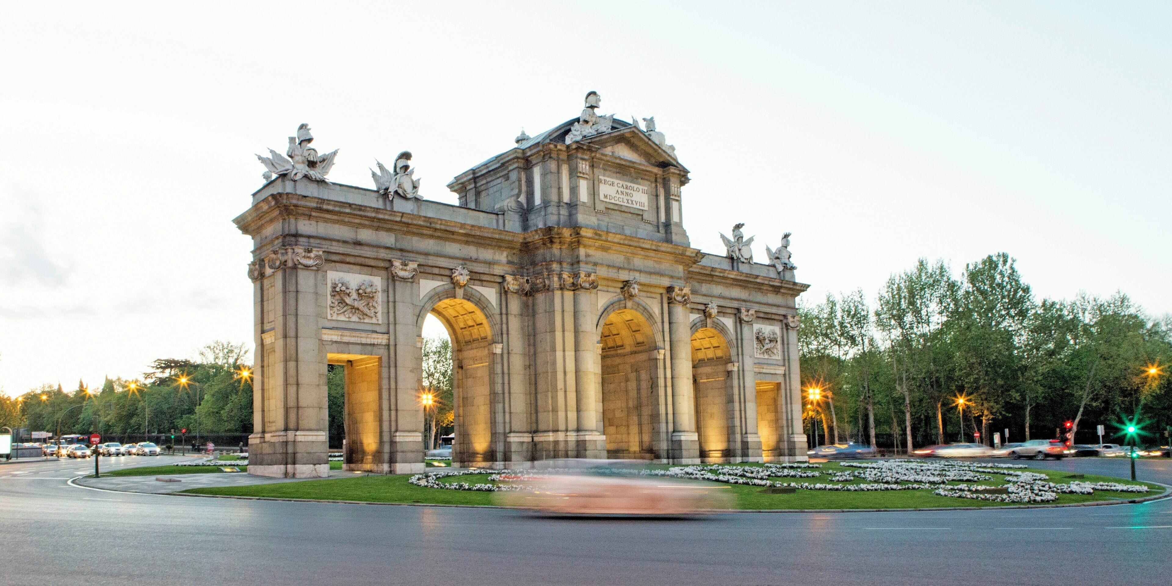 A grand stone archway with statues on top, surrounded by a well-maintained garden and a busy road in the background.