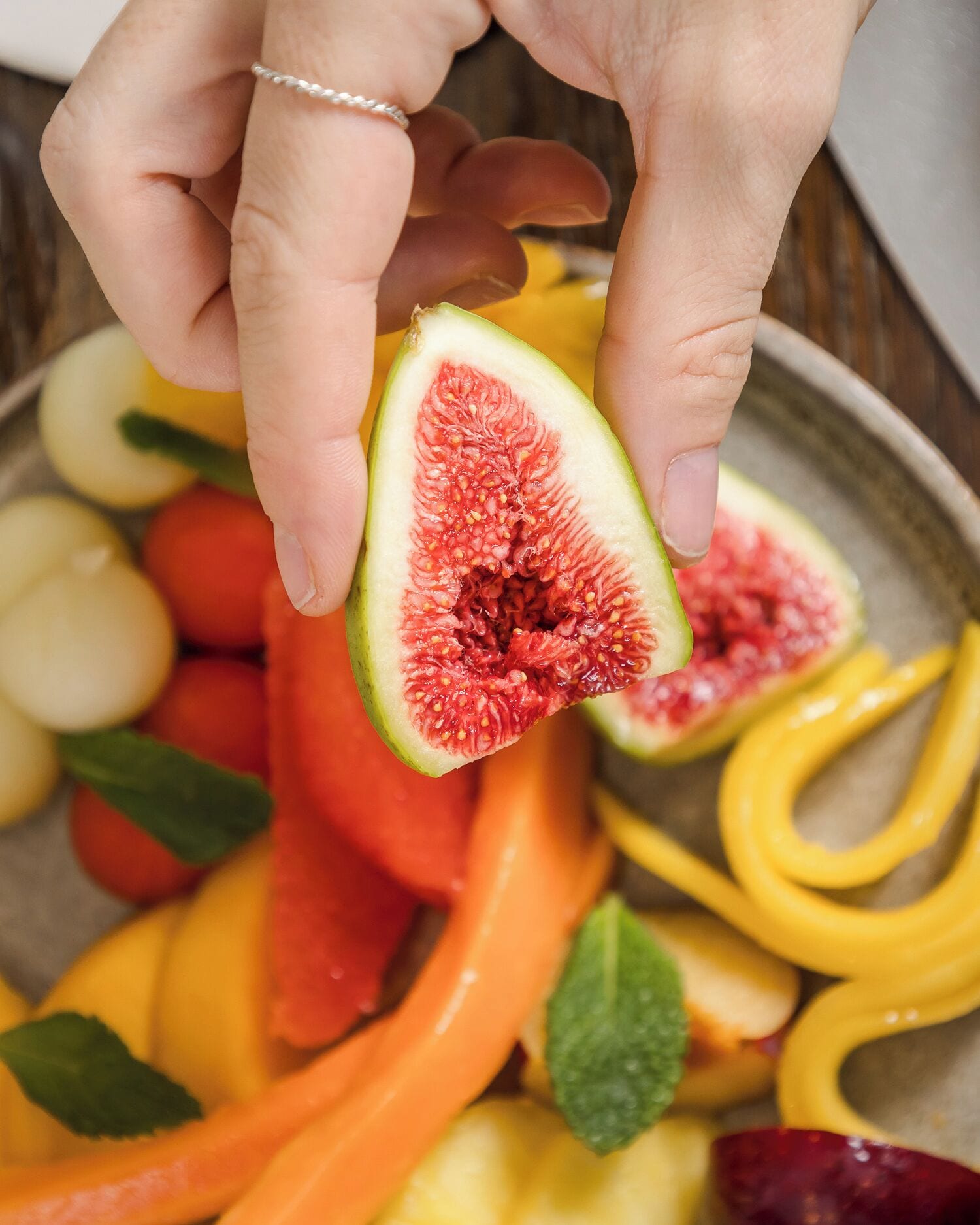 Una persona sostiene una higo cortada por la mitad, mostrando su interior con semillas, rodeada de diversas frutas y verduras.