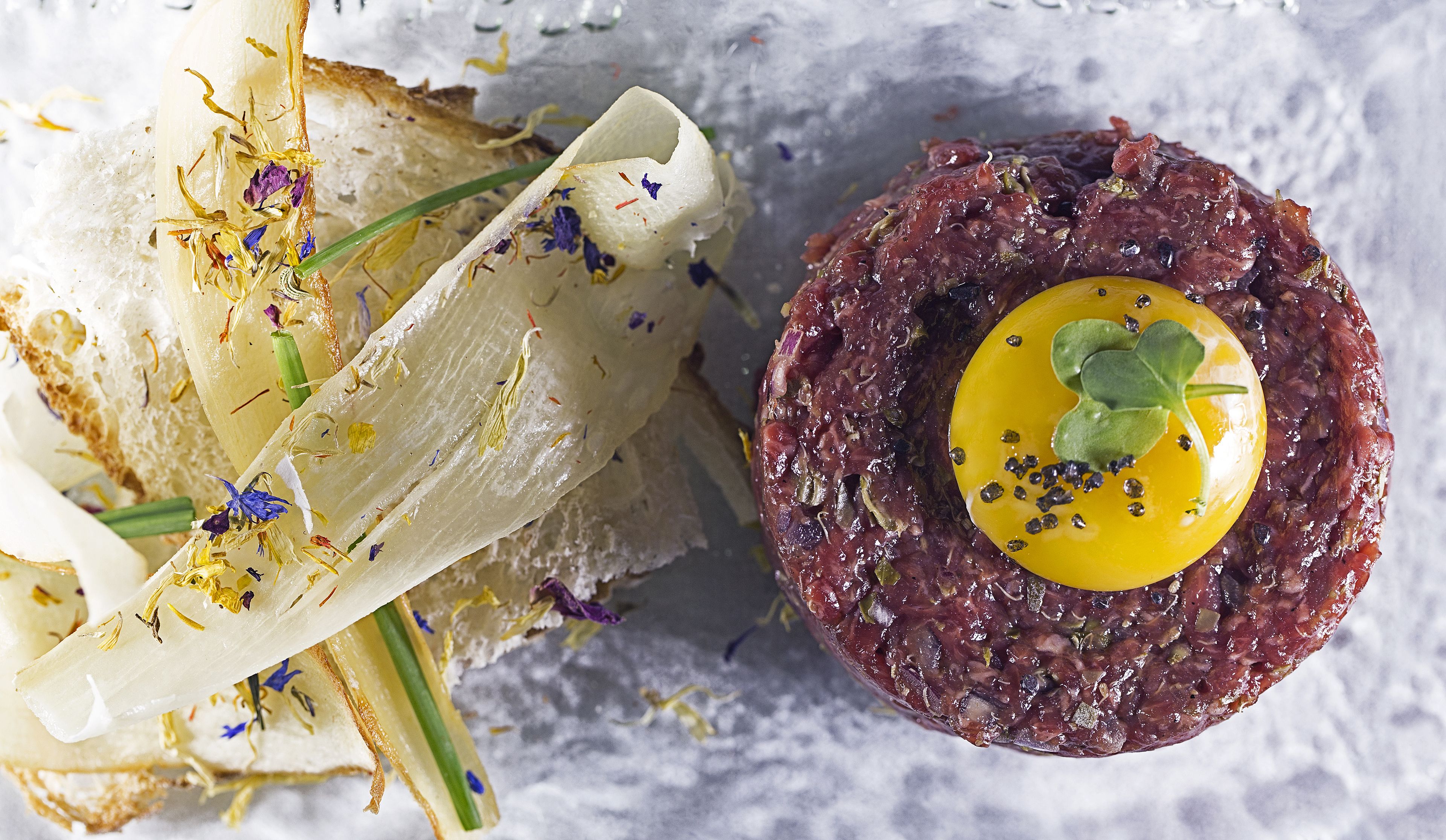 Steak Tartare mit rohem Eigelb und Garnitur, dazu eine Kräuterbrot Scheibe.