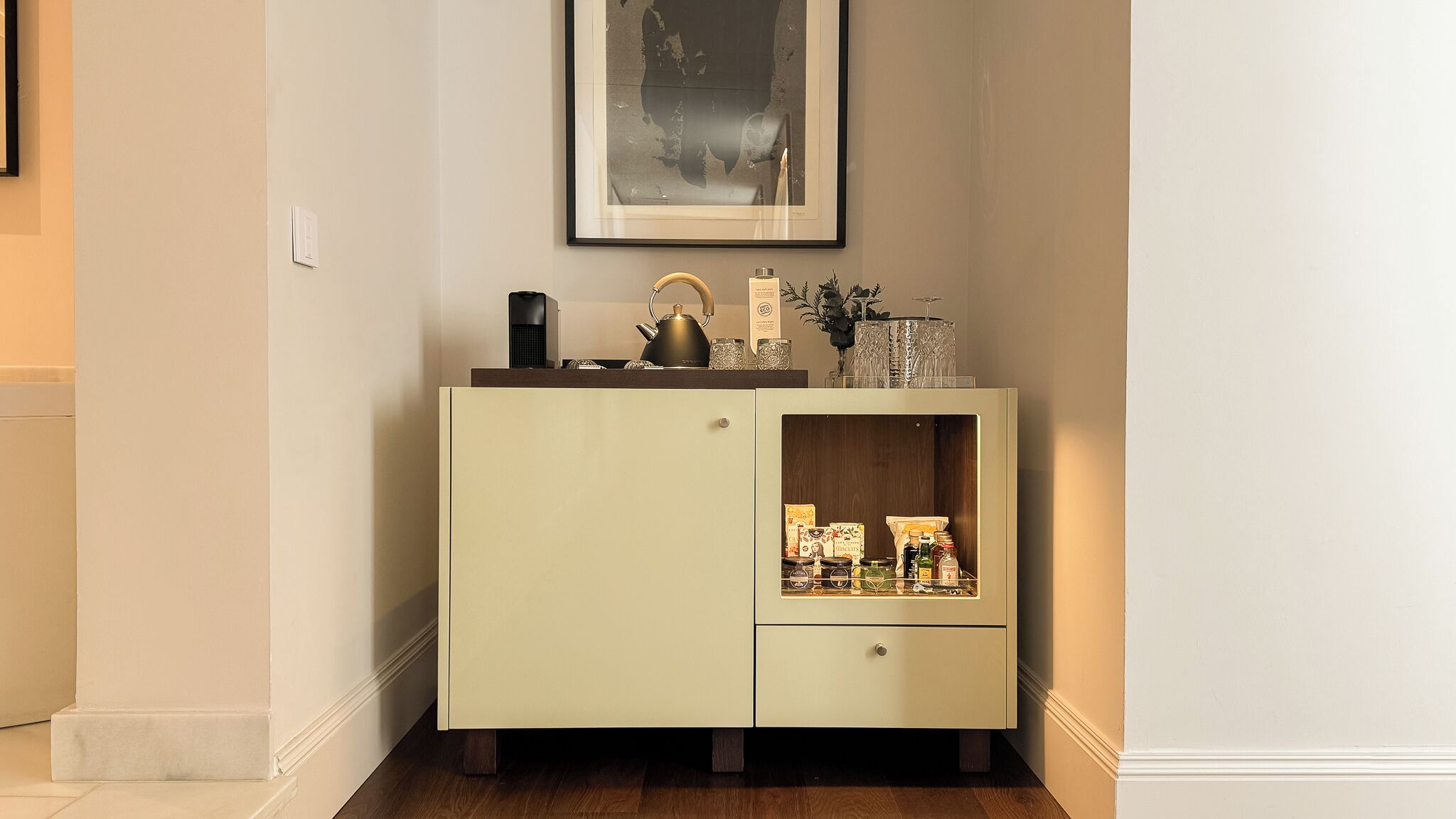 A corner cabinet with a tea kettle, glassware, and a framed picture on top.