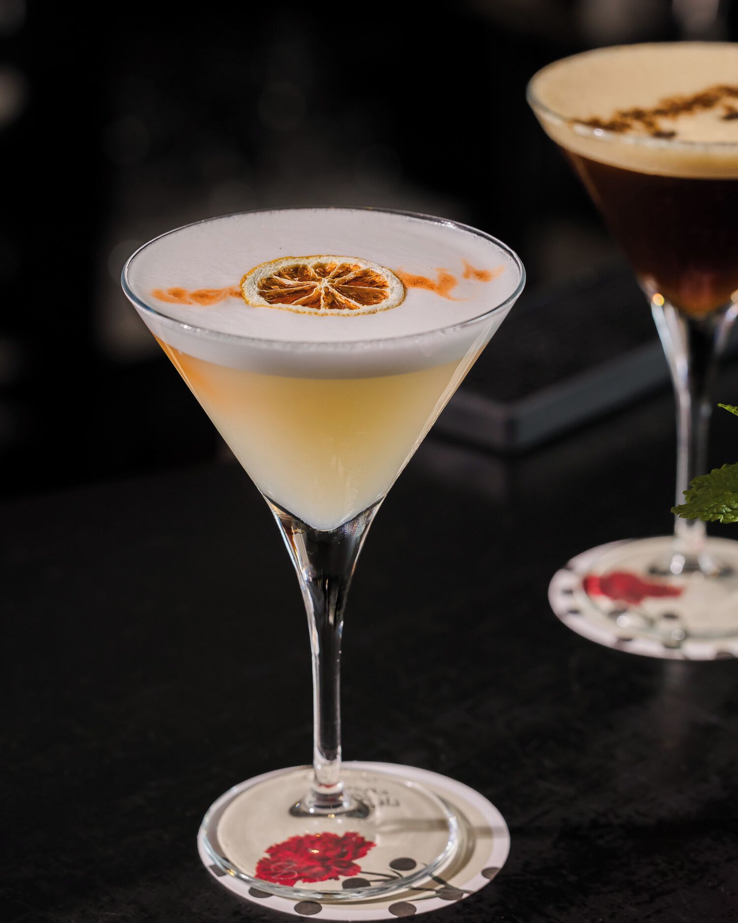 Un verre à martini contenant une boisson à plusieurs couches, décoré d’une tranche d’orange séchée, posé sur un dessous de verre orné d’une fleur rouge.