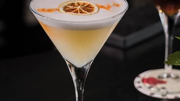Un verre à martini contenant une boisson à plusieurs couches, décoré d’une tranche d’orange séchée, posé sur un dessous de verre orné d’une fleur rouge.