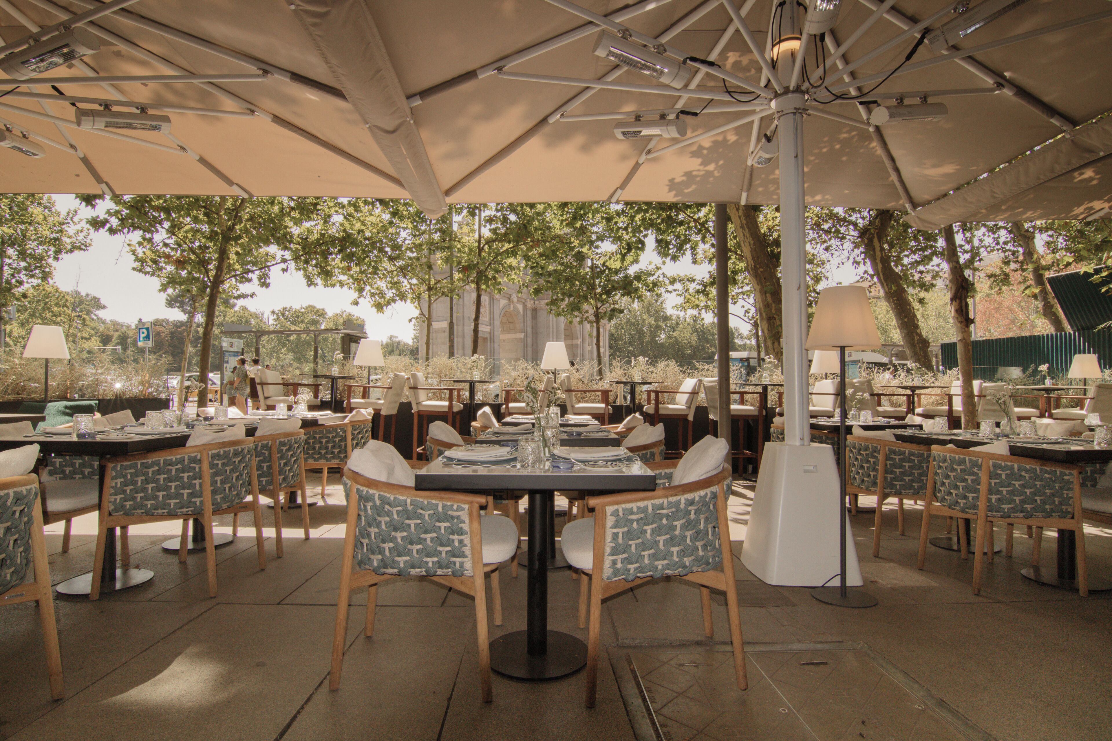 An outdoor dining area with multiple tables and chairs, umbrellas, and a view of trees and buildings in the background.