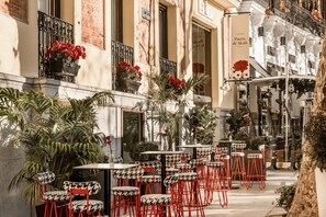 Ein Straßencafé mit roten und schwarzen Stuhl- und Tischsets, umgeben von Topfpflanzen und einer Ziegelhausfassade.