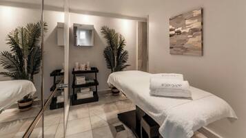 A spa room with two treatment beds, a mirror, a shelf with toiletries, and a painting on the wall.