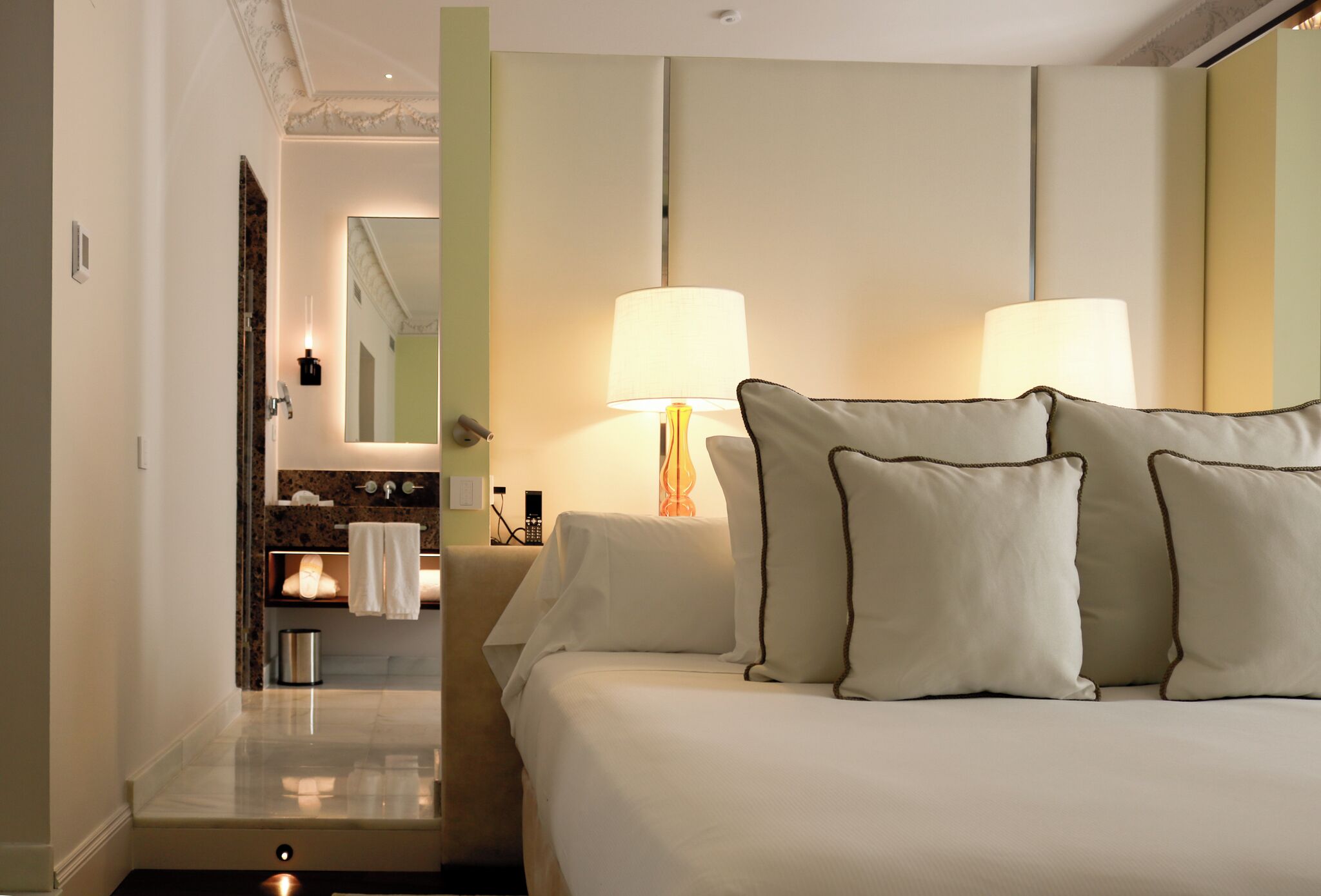 A hotel room with a bed, two bedside lamps, a nightstand, a mirror, a bathroom with a sink and towel rack, and a door leading to another room.