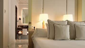A hotel room with a bed, two bedside lamps, a nightstand, a mirror, a bathroom with a sink and towel rack, and a door leading to another room.