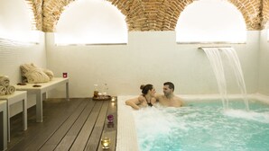 Una pareja en un jacuzzi con cascada, rodeada por una terraza de madera y una pared de azulejos con un arco de ladrillo.
