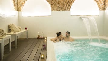 A couple in a hot tub with a waterfall feature, surrounded by a wooden deck and a tiled wall with a brick arch.