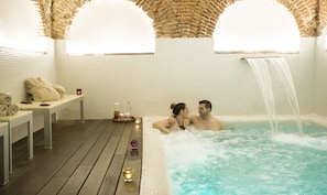 A couple in a hot tub with a waterfall feature, surrounded by a wooden deck and a tiled wall with a brick arch.