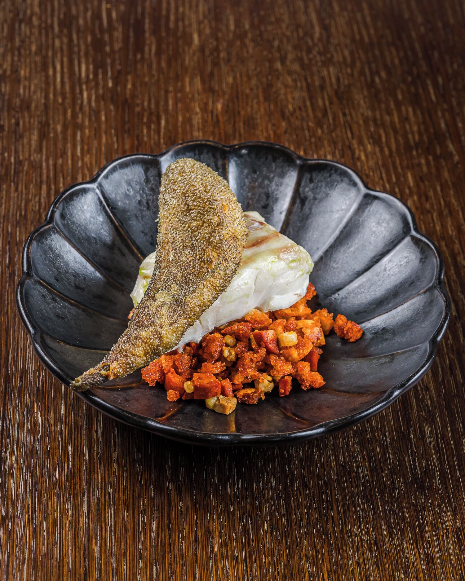 A plate with a fish, a piece of bread, and a side of diced vegetables on a dark, scalloped plate.