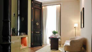 Una habitación con una puerta de madera oscura, un sillón color crema, una mesita de noche con una lámpara y una ventana con cortinas.