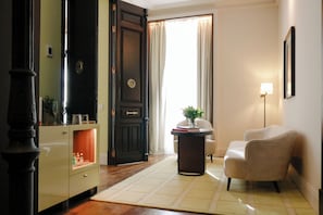 Une pièce avec une porte en bois foncé, un fauteuil beige, une petite table avec une lampe et une fenêtre avec des rideaux.