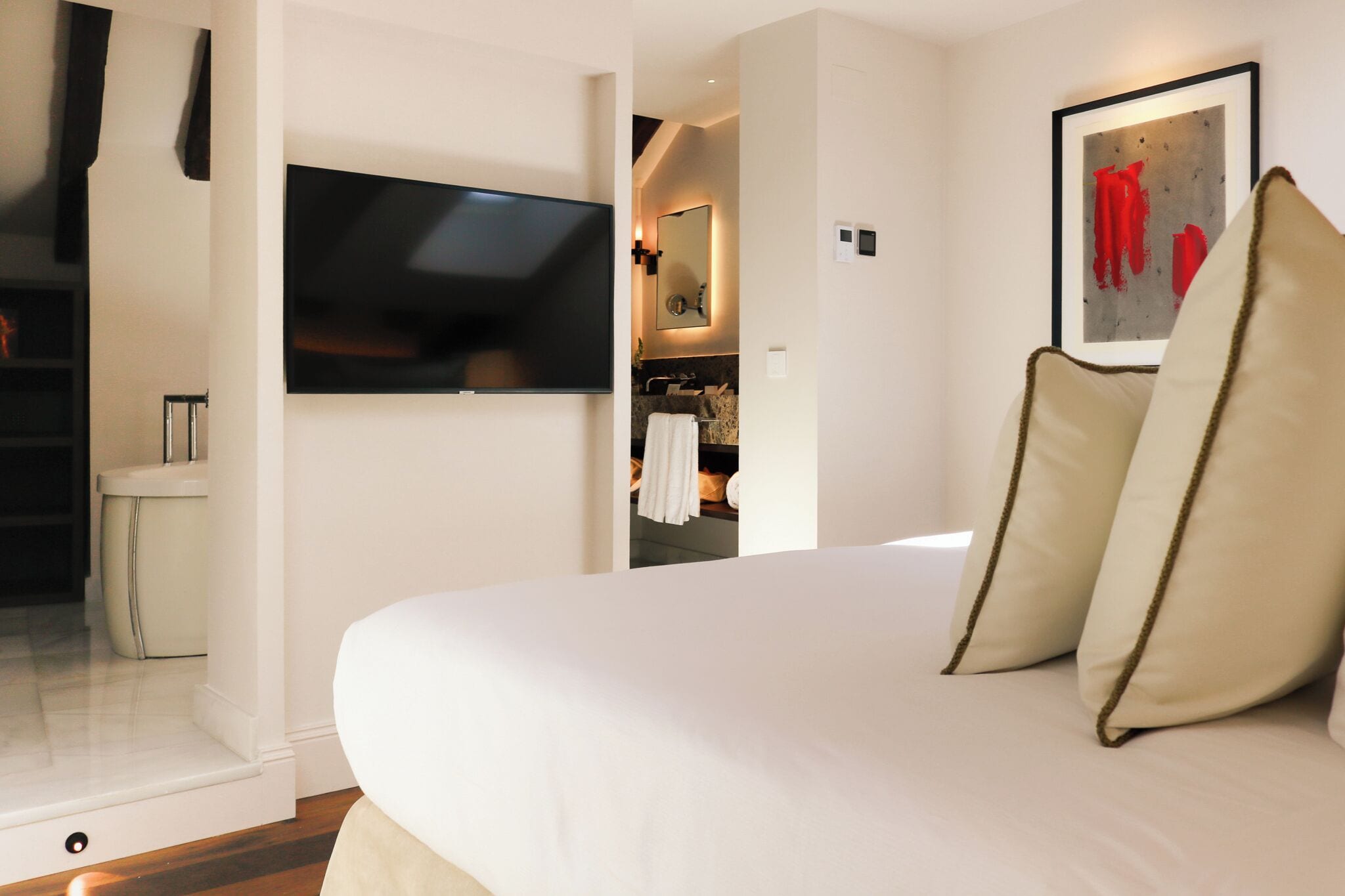 Quarto de hotel com uma cama grande, televisão presa à parede, banheiro visível por uma porta aberta e uma obra de arte emoldurada na parede.