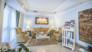 A well-lit living room with patterned sofas, a coffee table, and a bookshelf filled with books and magazines. - Hotel Villa Daphne (Giardini Naxos)