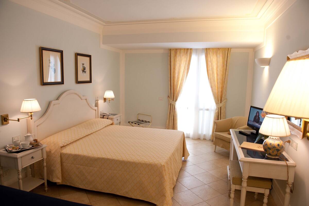 Ein Hotelzimmer mit Bett, Nachttischen, einem Schreibtisch mit Lampe, einem Sessel und einem Fernseher, sowie Vorhängen.