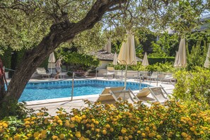 Un área de piscina con sillas de descanso y sombrillas, rodeada de vegetación y un árbol grande.