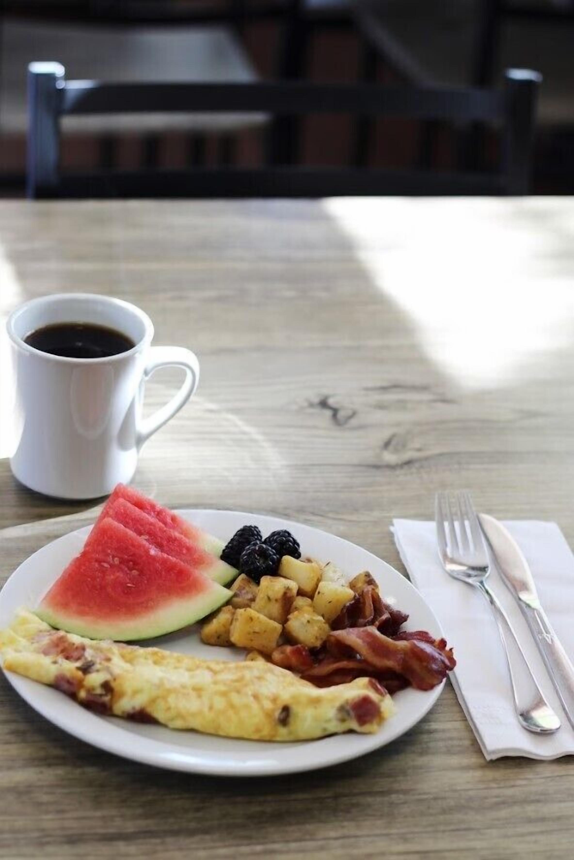 Une assiette de petit-déjeuner comprenant une omelette, de la pastèque, des mûres, des pommes de terre et du bacon, accompagnée d’une tasse de café sur une table en bois.