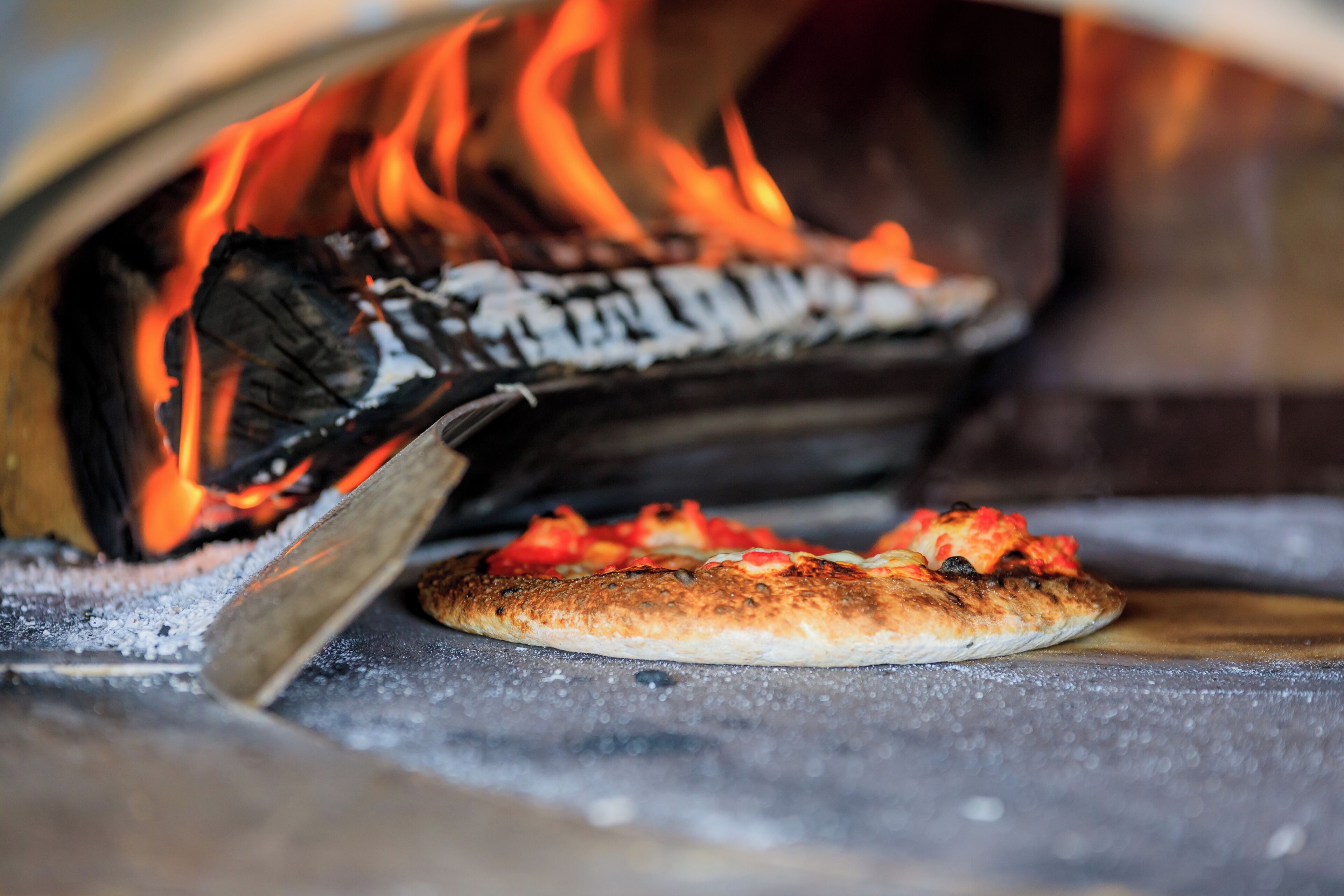 A pizza oven with a pizza inside, flames visible, and a pizza peel in front.