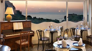 A dining area with a piano, tables set for two, and a view of the sea.