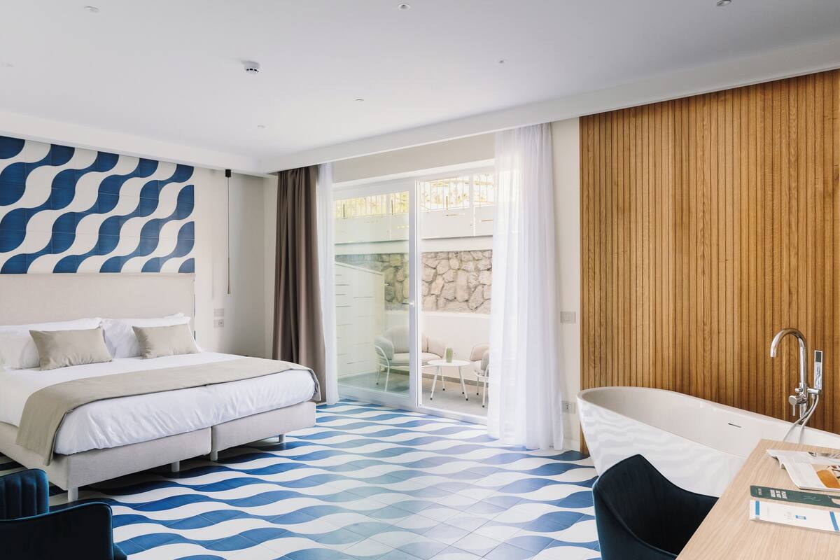 A modern hotel room with a bed, a bathtub, a dining table, and a view of the outdoor area.
