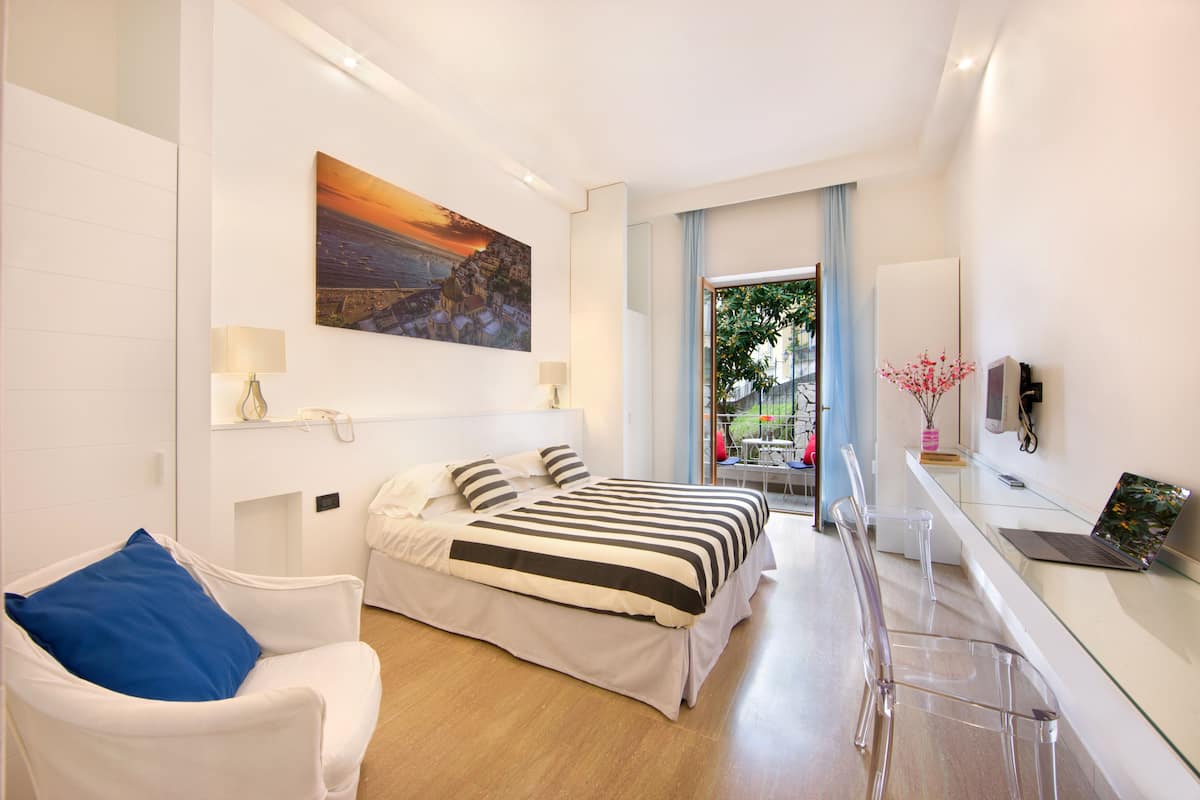 A modern hotel room with a bed, a desk with a laptop, a chair, and a painting on the wall.