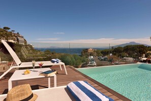 Un espace aménagé au bord de la piscine, avec des chaises longues, une table où sont servis des boissons et une vue sur un paysage côtier.