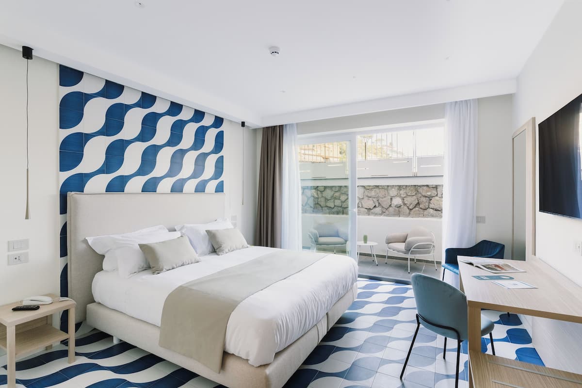 A modern hotel room with a large bed, a desk, and a balcony with seating.