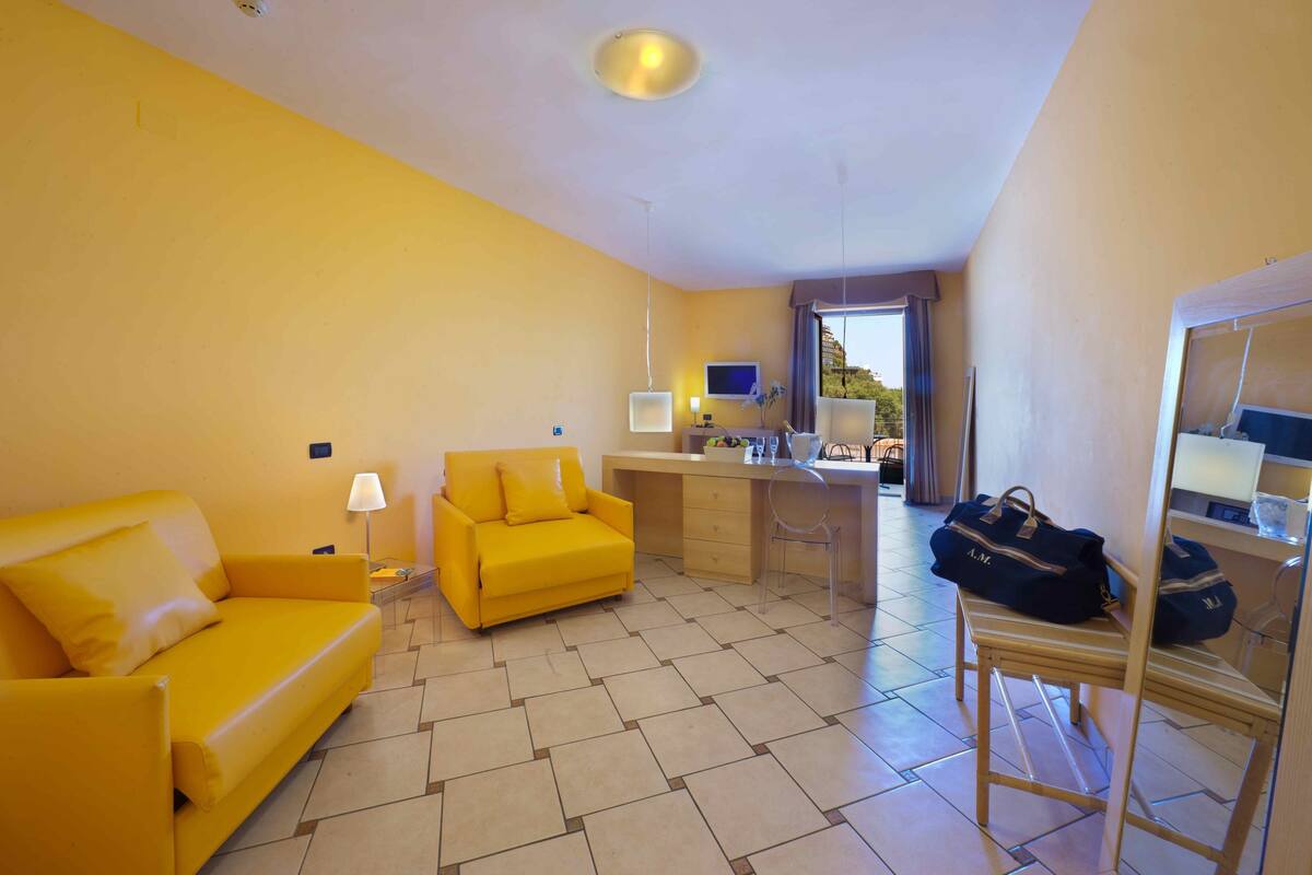 A compact hotel room with a yellow sofa, a small desk with a computer, a chair, and a suitcase on a stand.