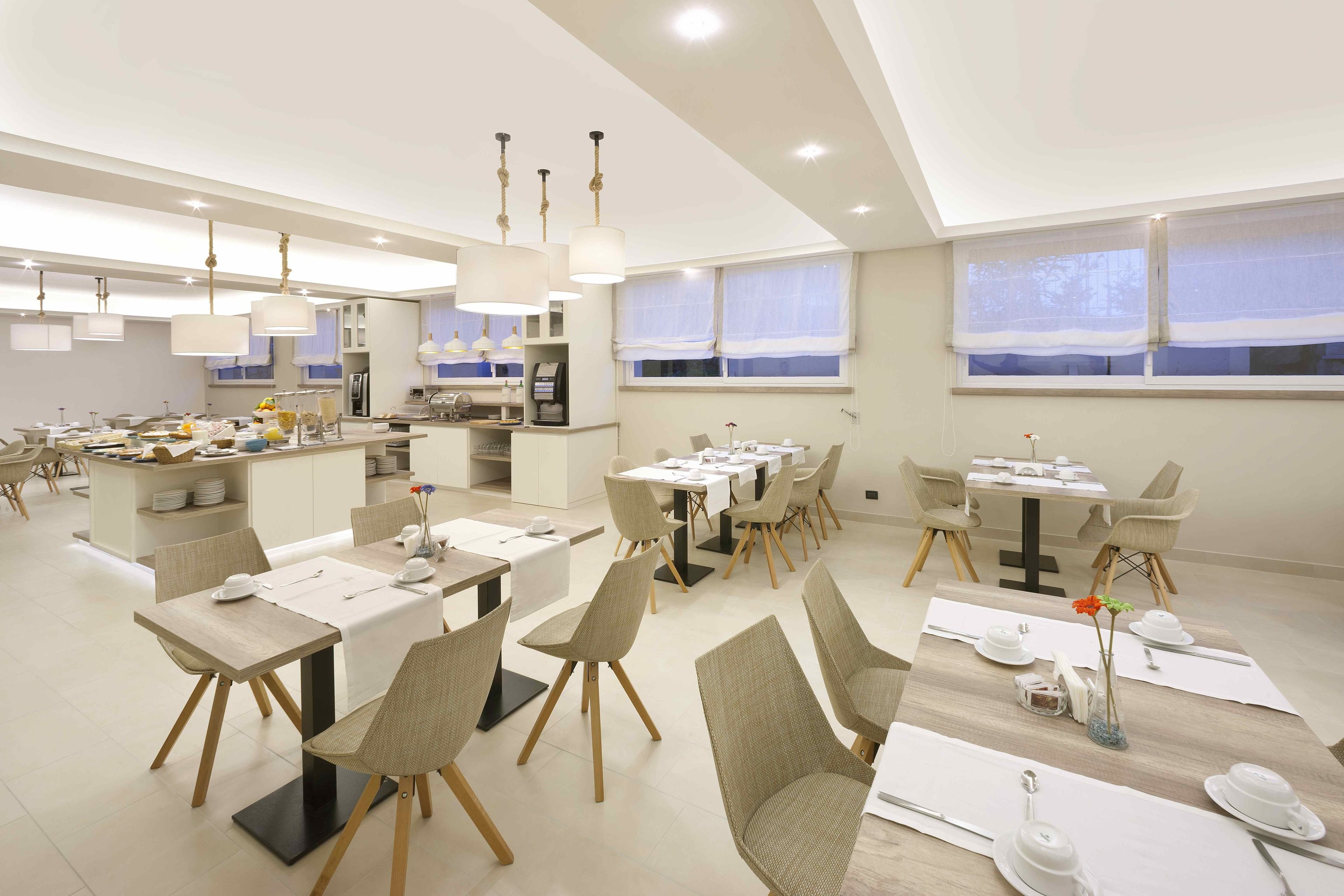 Un comedor moderno con varias mesas y sillas, una cocina al fondo y amplias ventanas con cortinas ligeras.