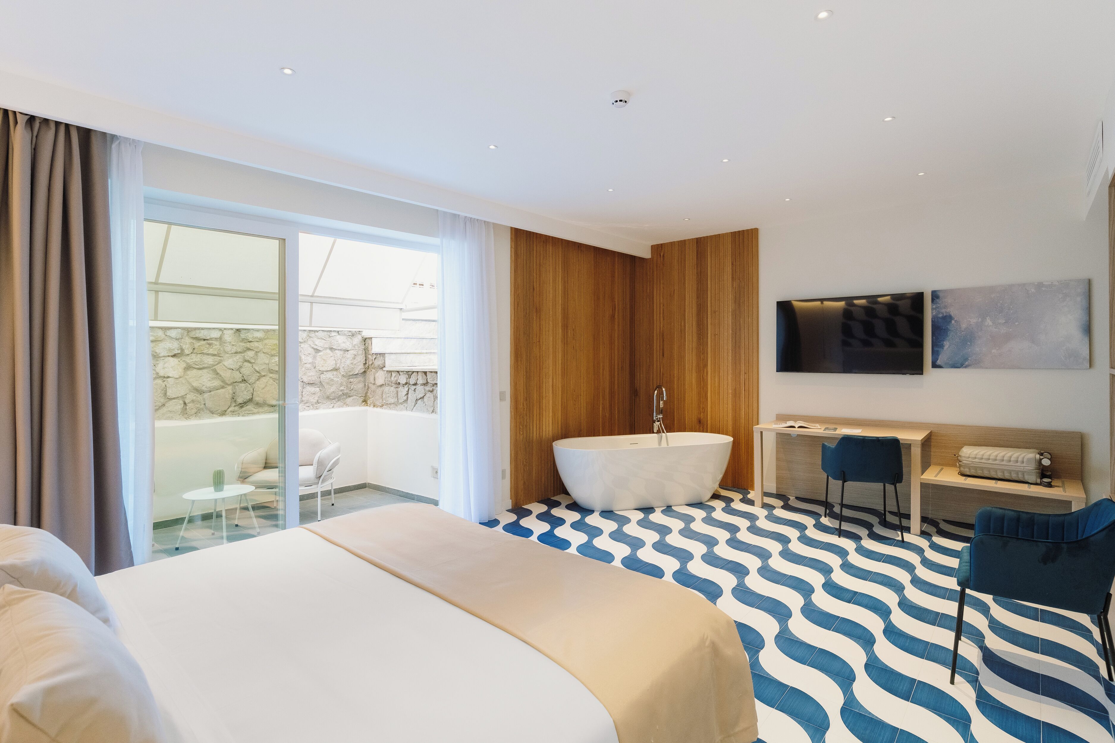 A modern hotel room with a large bed, a bathroom with a freestanding bathtub, a desk, and a TV mounted on the wall.
