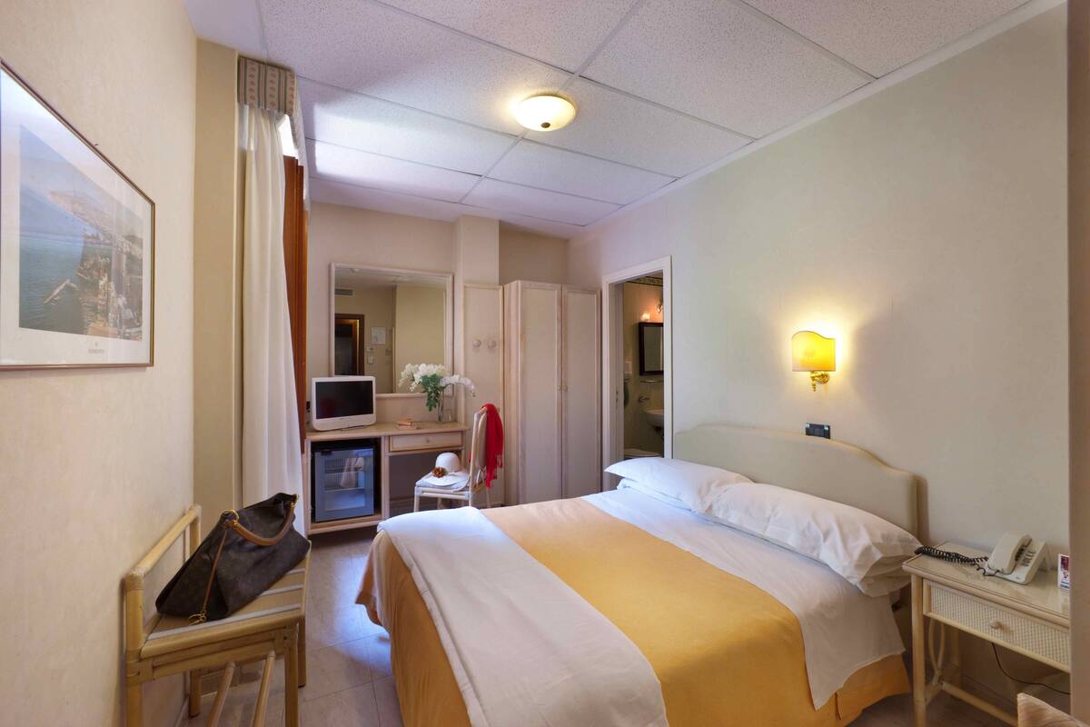 A hotel room with a bed, a desk with a computer, a chair, a handbag, and a telephone.