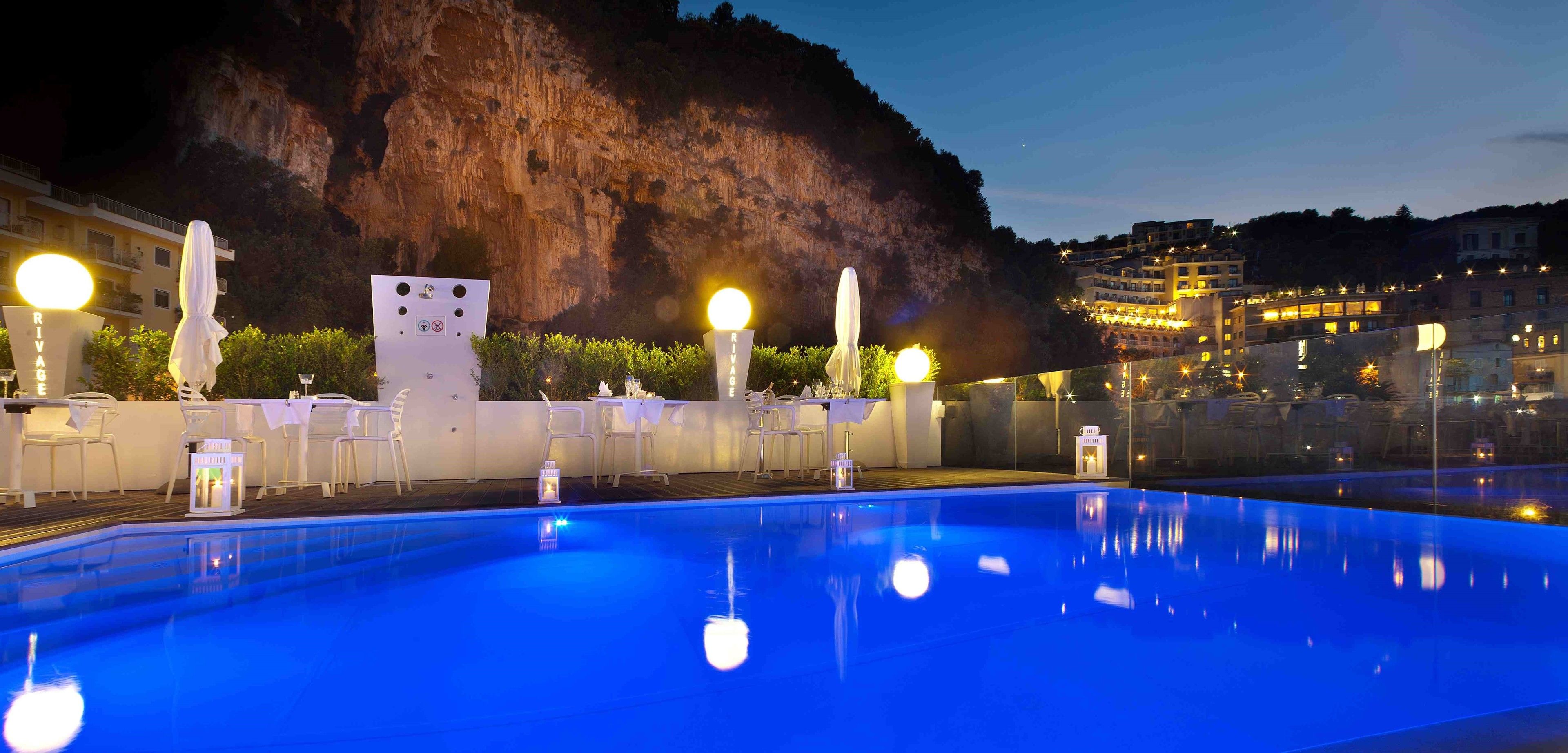 A swimming pool with lounge chairs and umbrellas, a sign reading "RIVAGE", and a mountainous backdrop.