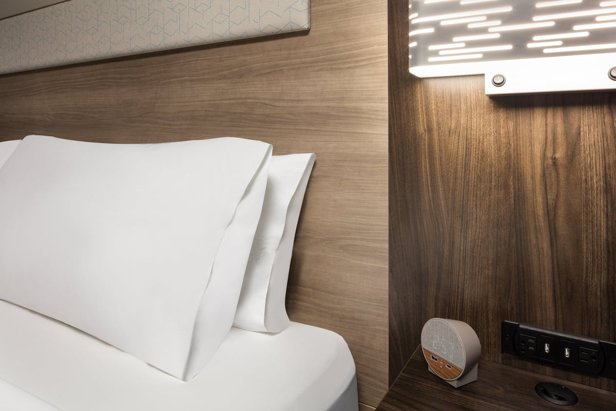 A neatly made bed with white linens, a wall-mounted light panel, and a speaker on the floor.