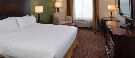Ein Hotelzimmer mit einem großen Bett, einem Schreibtisch mit Computer, einem Sessel, einem Fernseher und einem Fenster mit Vorhängen.