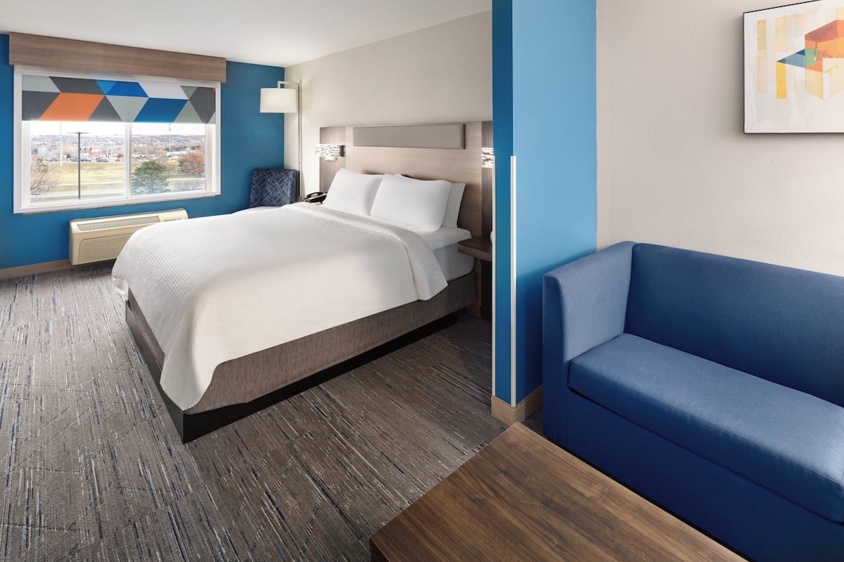A modern hotel room with a large bed, a blue sofa, a view of the city through the window, and a geometric-patterned curtain.