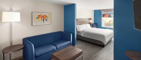 A hotel room with a blue sofa, a wooden coffee table, a bed with white bedding, and a painting on the wall.