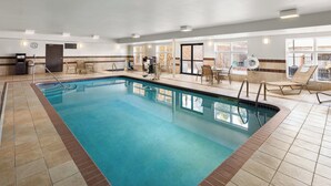 An indoor swimming pool with clear blue water, surrounded by lounge chairs and a sunbed, with a view of trees through large windows.